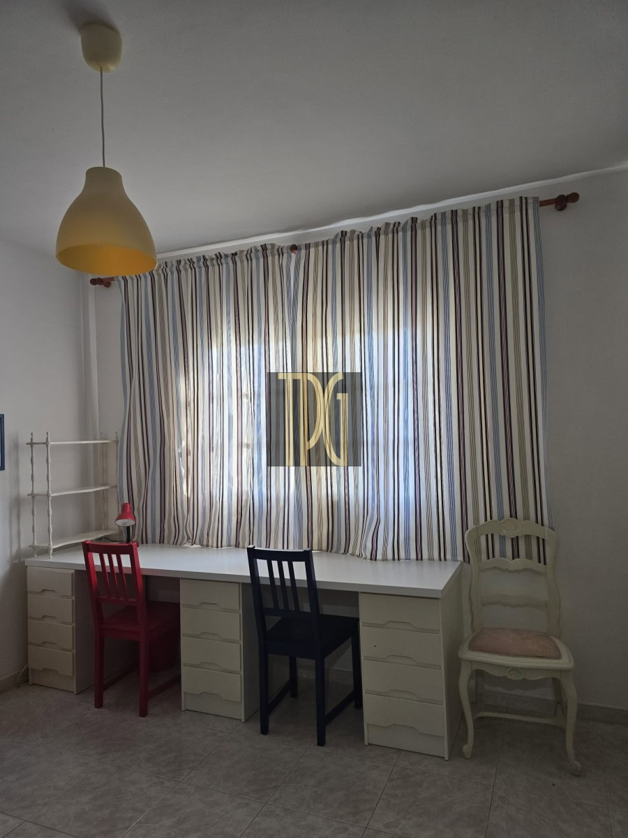 White desk with multiple drawers runs along a window dressed in striped curtains; red, dark blue, and white ornate chairs sit in front (lamp overhead).
