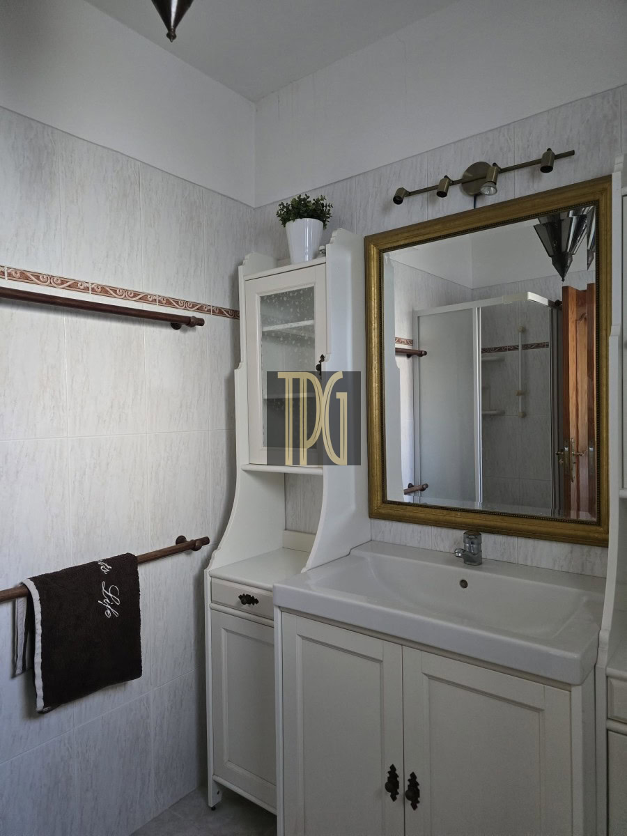 White bathroom vanity with sink, storage cabinets, and a gold-framed mirror; a small white cabinet with a glass door sits beside it, topped by a potted plant.