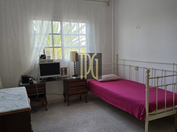 Light-filled bedroom with a white metal-framed single bed in pink sheets, a marble-top dresser, and a desk with a computer near a window.