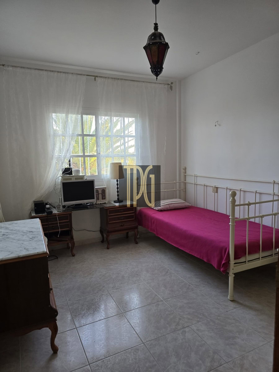Light-filled bedroom with a white metal-framed single bed in pink sheets, a marble-top dresser, and a desk with a computer near a window.