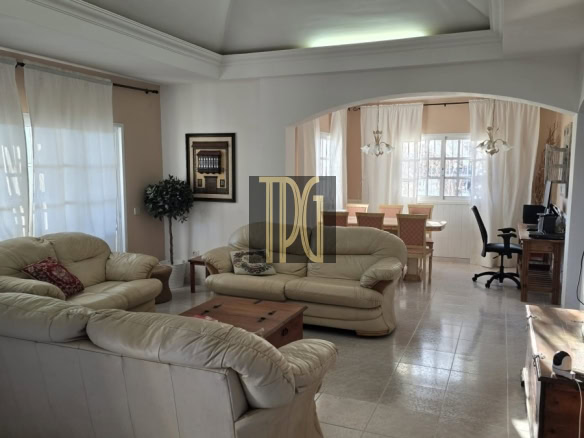 Bright open-plan living room with beige leather sofas, glass coffee table, and an arch opening to a dining area with a long table and chairs.
