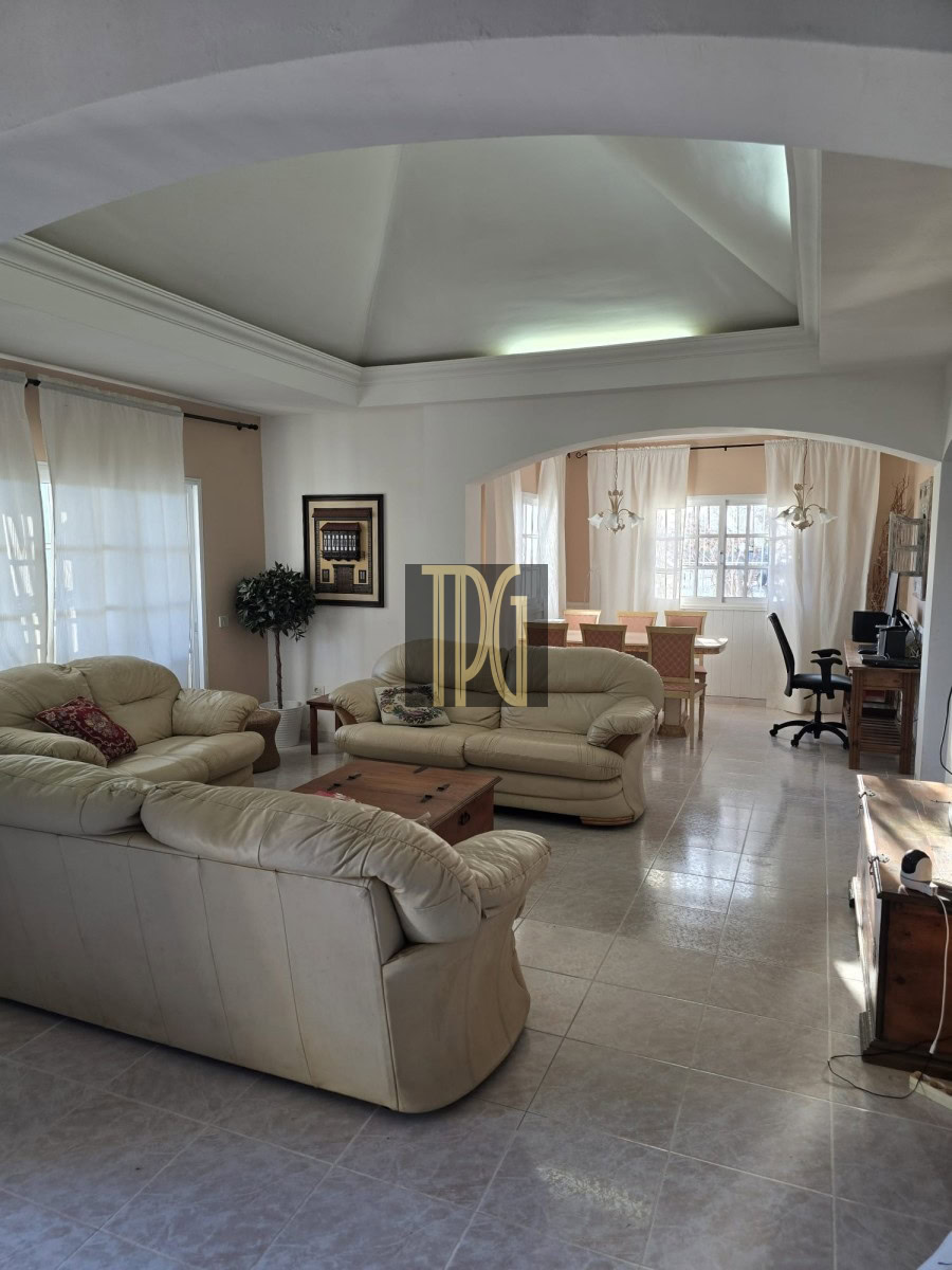 Bright open-plan living room with beige leather sofas, glass coffee table, and an arch opening to a dining area with a long table and chairs.