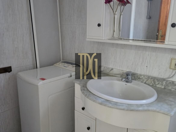 Bathroom with a white top-loading washing machine beside a marble countertop sink and mirror cabinet, with purple flowers in vases.