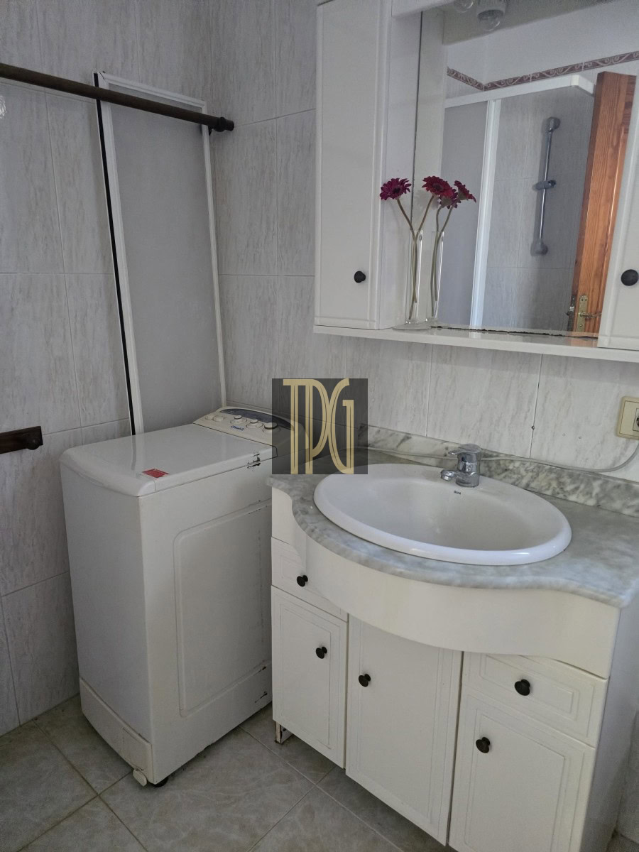 Bathroom with a white top-loading washing machine beside a marble countertop sink and mirror cabinet, with purple flowers in vases.