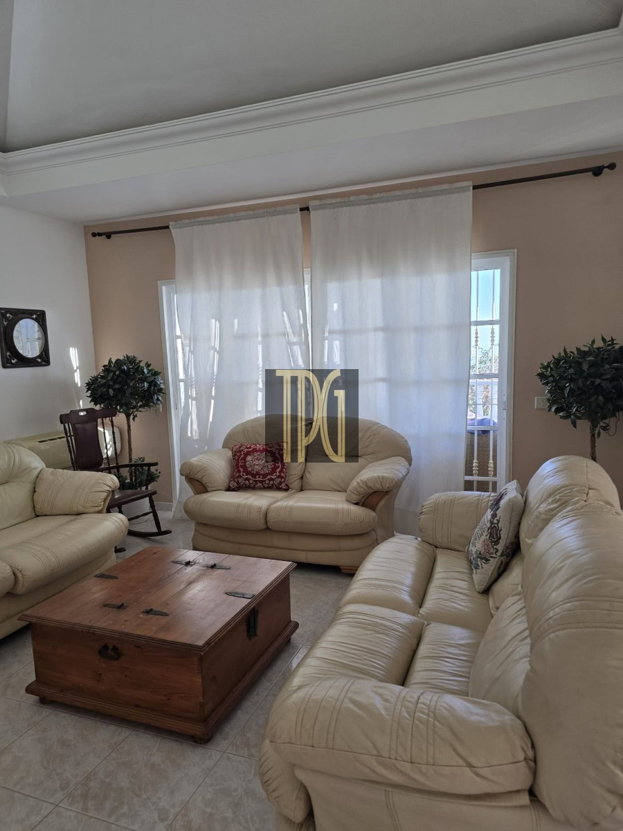 Beige leather living room with matching sofa set, a wooden trunk-style coffee table, and sheer white curtains with potted plants nearby.