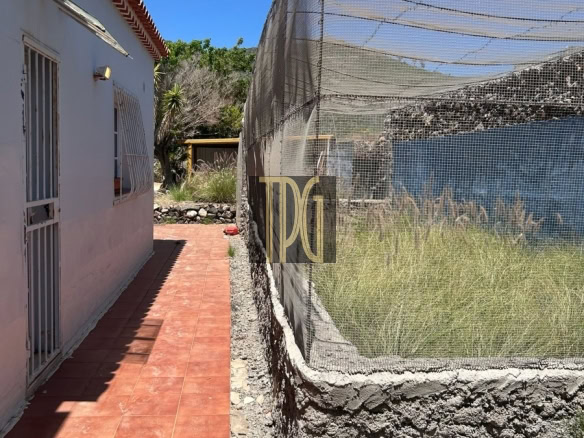 Narrow outdoor path between a white building with a red-tiled roof and a mesh-enclosed fenced area under a blue sky.