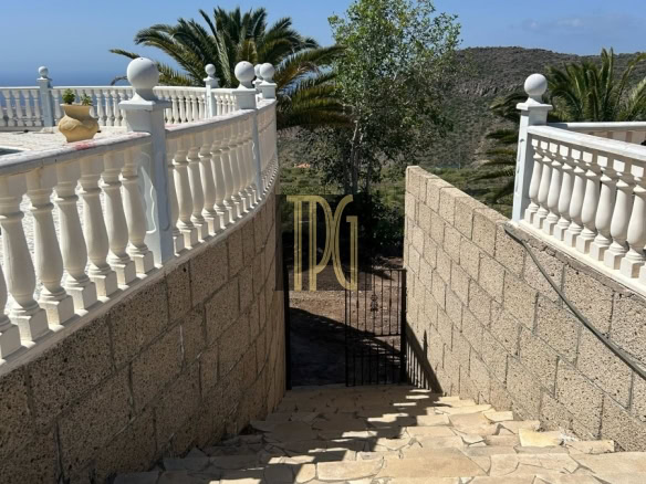 Stone stairway descending between tan stone walls with white balustrades, leading to a gate amid palm trees and hills under a blue sky