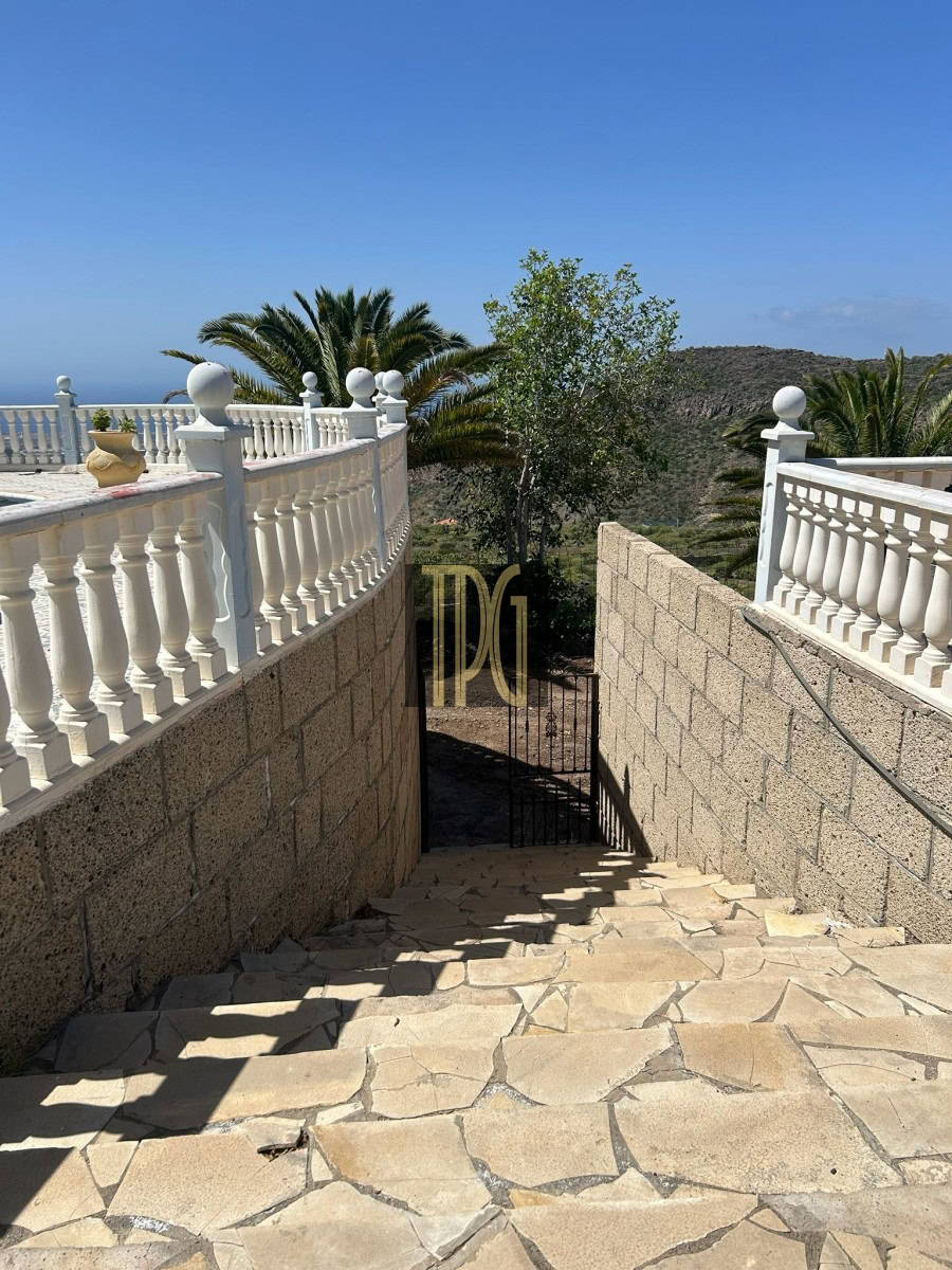 Stone stairway descending between tan stone walls with white balustrades, leading to a gate amid palm trees and hills under a blue sky