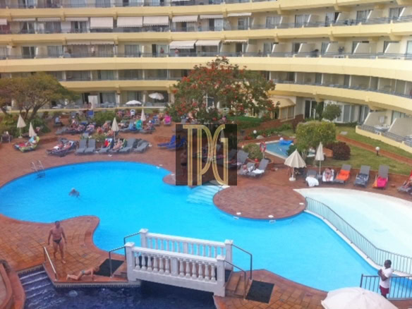 Hotel pool courtyard with sun loungers, umbrellas, and guests relaxing by a curved blue pool surrounded by a beige building.
