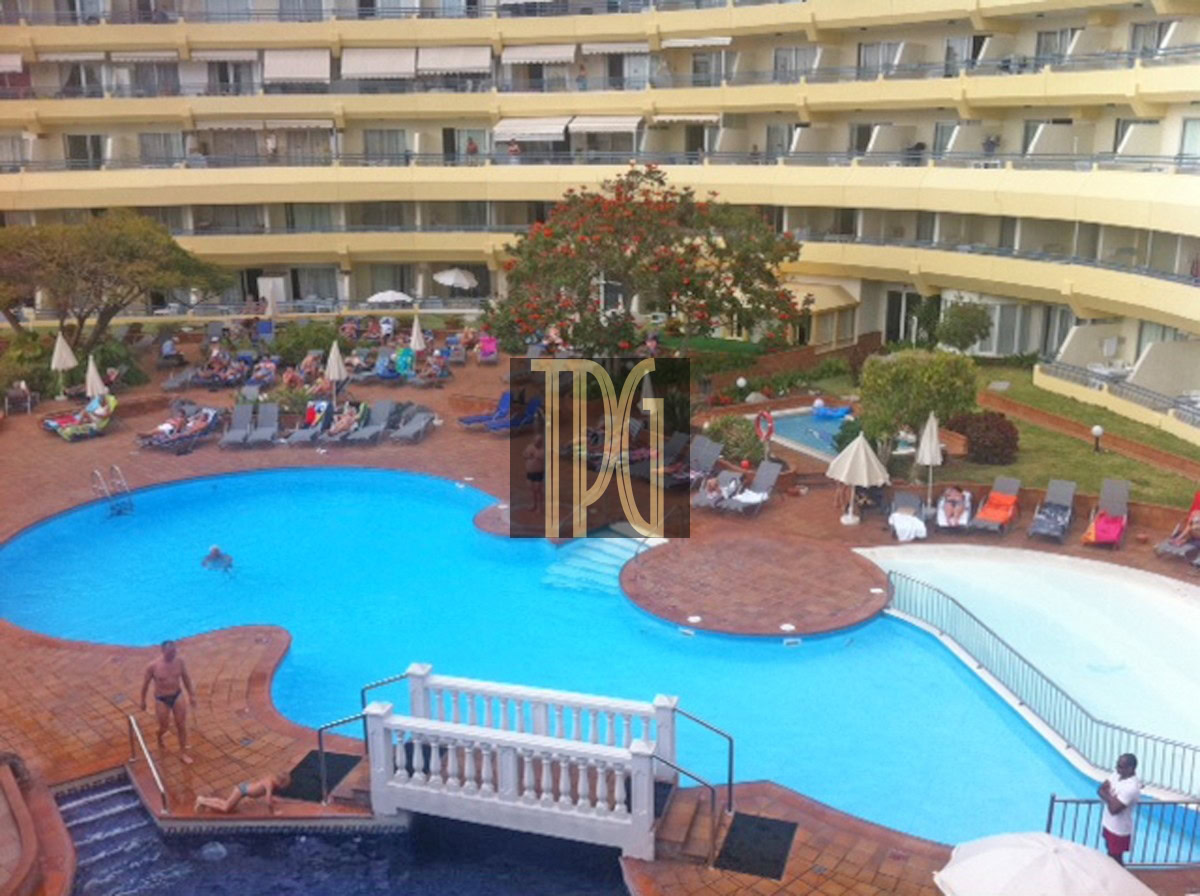 Hotel pool courtyard with sun loungers, umbrellas, and guests relaxing by a curved blue pool surrounded by a beige building.