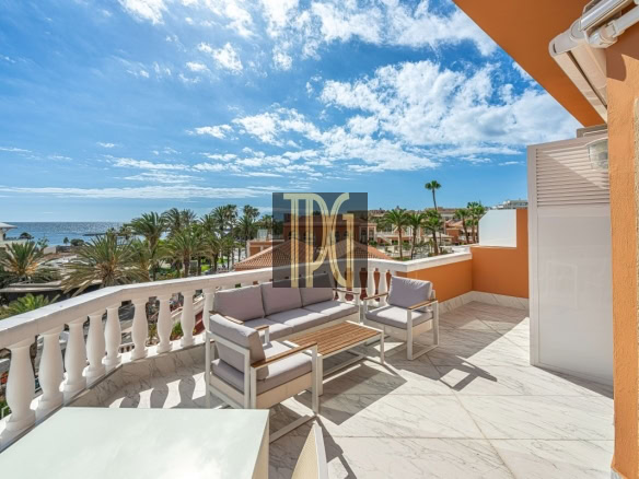 Sunny terrace with white marble floor and beige outdoor furniture overlooking a palm-lined street and the sea.