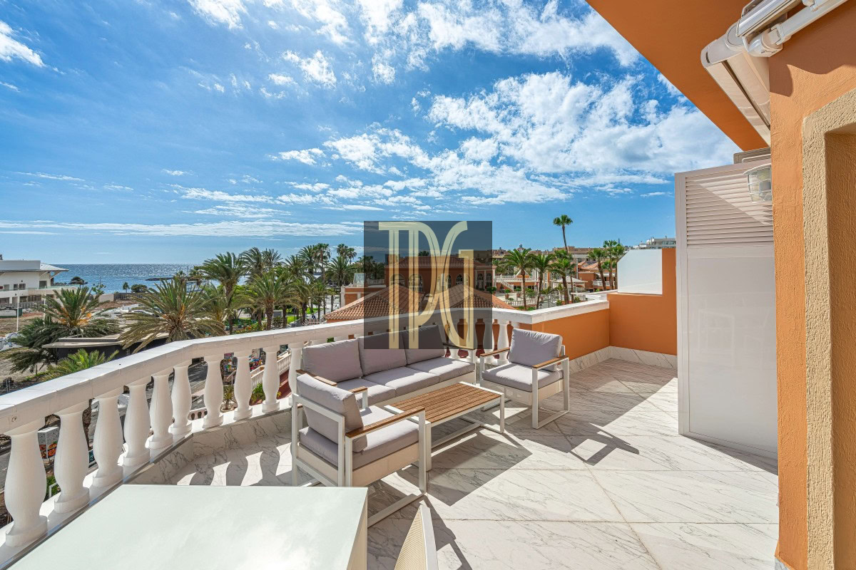 Sunny terrace with white marble floor and beige outdoor furniture overlooking a palm-lined street and the sea.