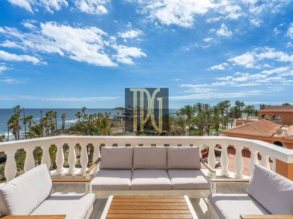 Outdoor balcony with cream cushions, white railings, and a wooden coffee table overlooking palm trees and the ocean under a blue sky.
