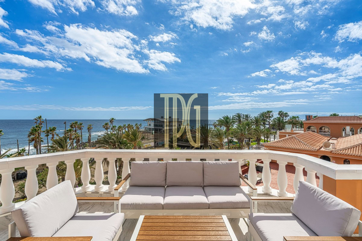 Outdoor balcony with cream cushions, white railings, and a wooden coffee table overlooking palm trees and the ocean under a blue sky.