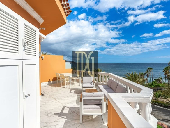 Outdoor terrace with white furniture overlooking the blue sea and palm trees on a sunny day, orange accent wall nearby.
