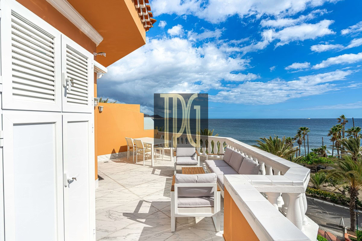 Outdoor terrace with white furniture overlooking the blue sea and palm trees on a sunny day, orange accent wall nearby.