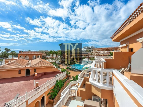 Resort complex with orange buildings, white railings, palm trees, and a central pool under a blue sky with clouds.