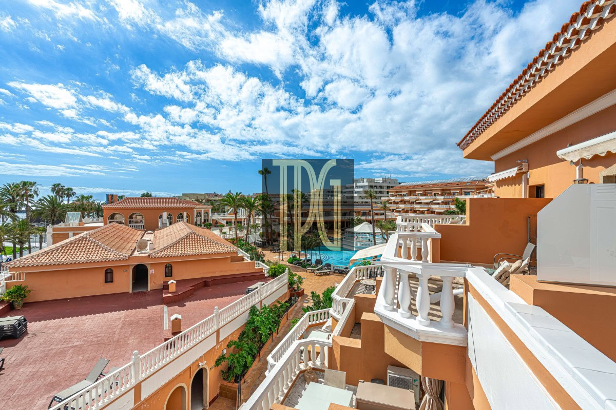Resort complex with orange buildings, white railings, palm trees, and a central pool under a blue sky with clouds.
