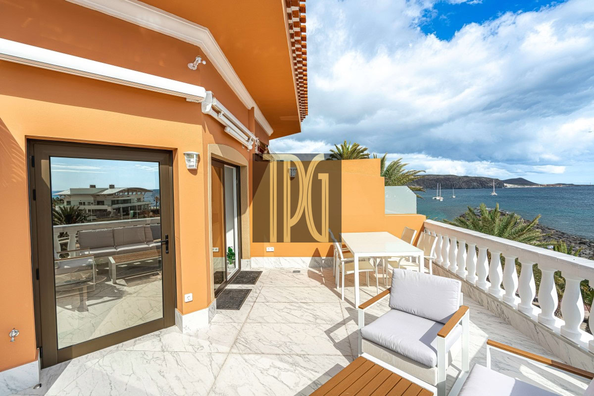 Orange-walled seaside terrace with white marble floor, outdoor seating, and a view of the ocean and palm trees.