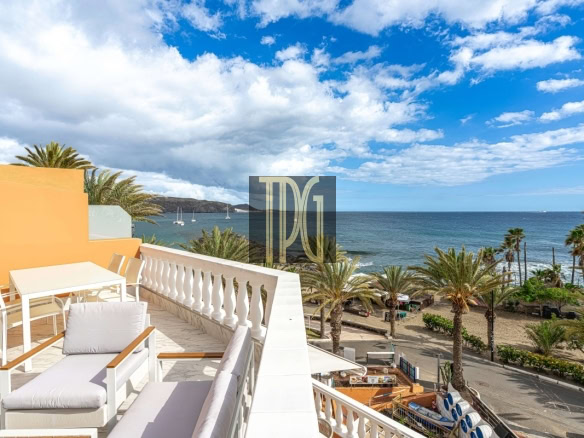 Sunny seaside terrace with white outdoor furniture, orange wall, and a balustrade overlooking a palm-lined beach and blue sea.
