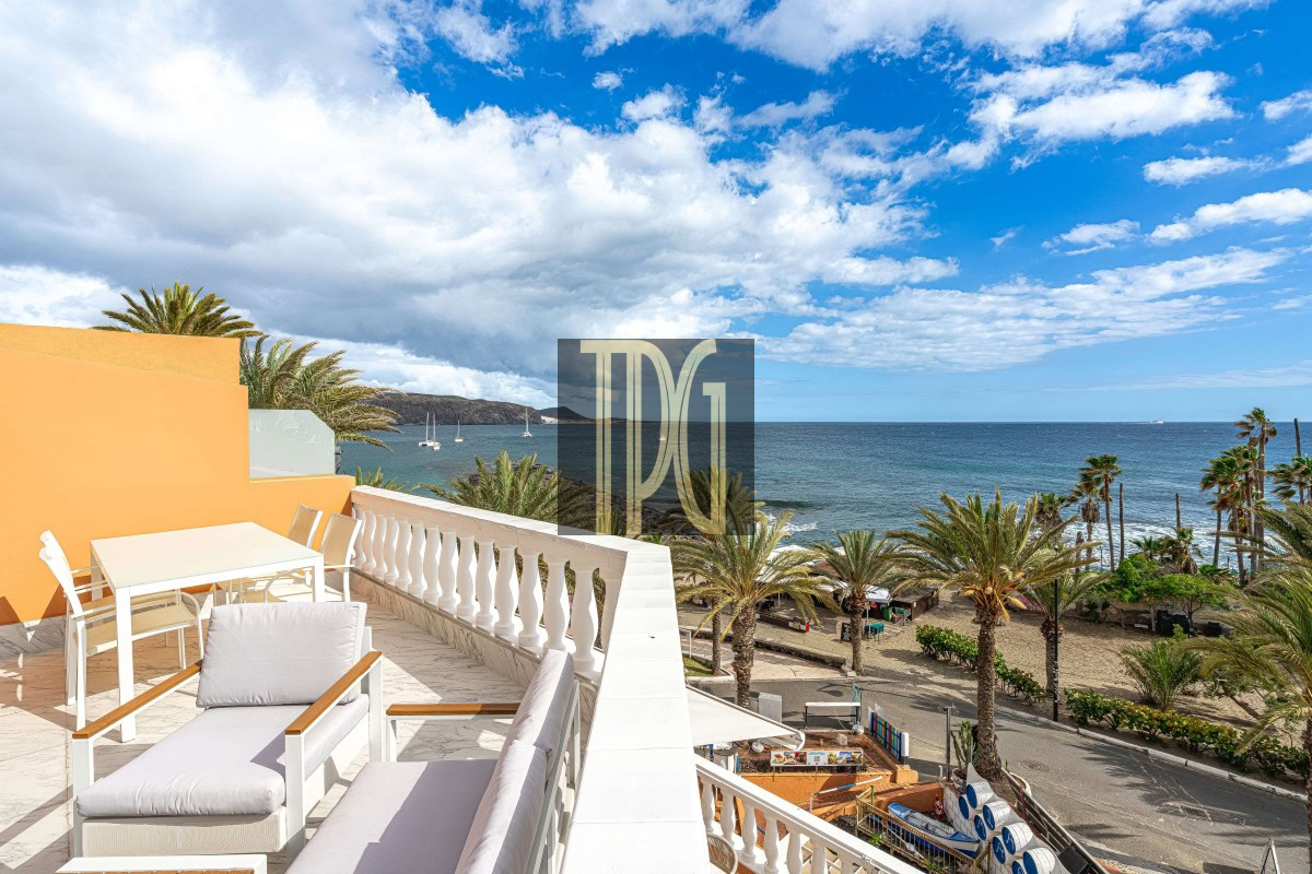 Sunny seaside terrace with white outdoor furniture, orange wall, and a balustrade overlooking a palm-lined beach and blue sea.