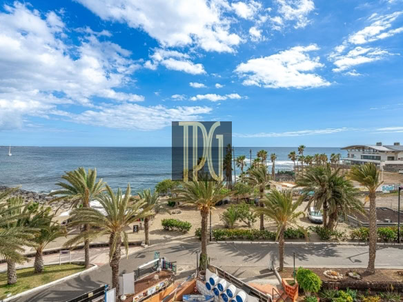 Coastal promenade lined with palm trees, overlooking a sandy beach and calm sea under a blue sky with scattered clouds.