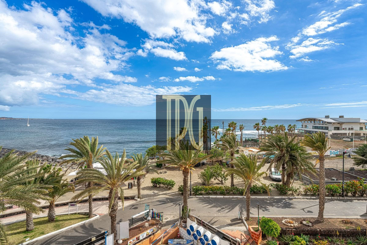 Coastal promenade lined with palm trees, overlooking a sandy beach and calm sea under a blue sky with scattered clouds.