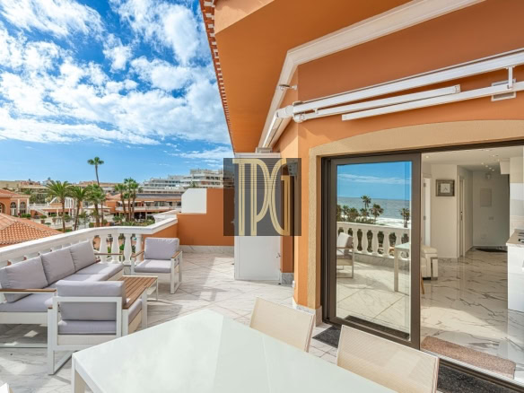Terrace with white outdoor seating, glass table, and ocean view; orange building exterior and sliding glass doors to the indoor kitchen.