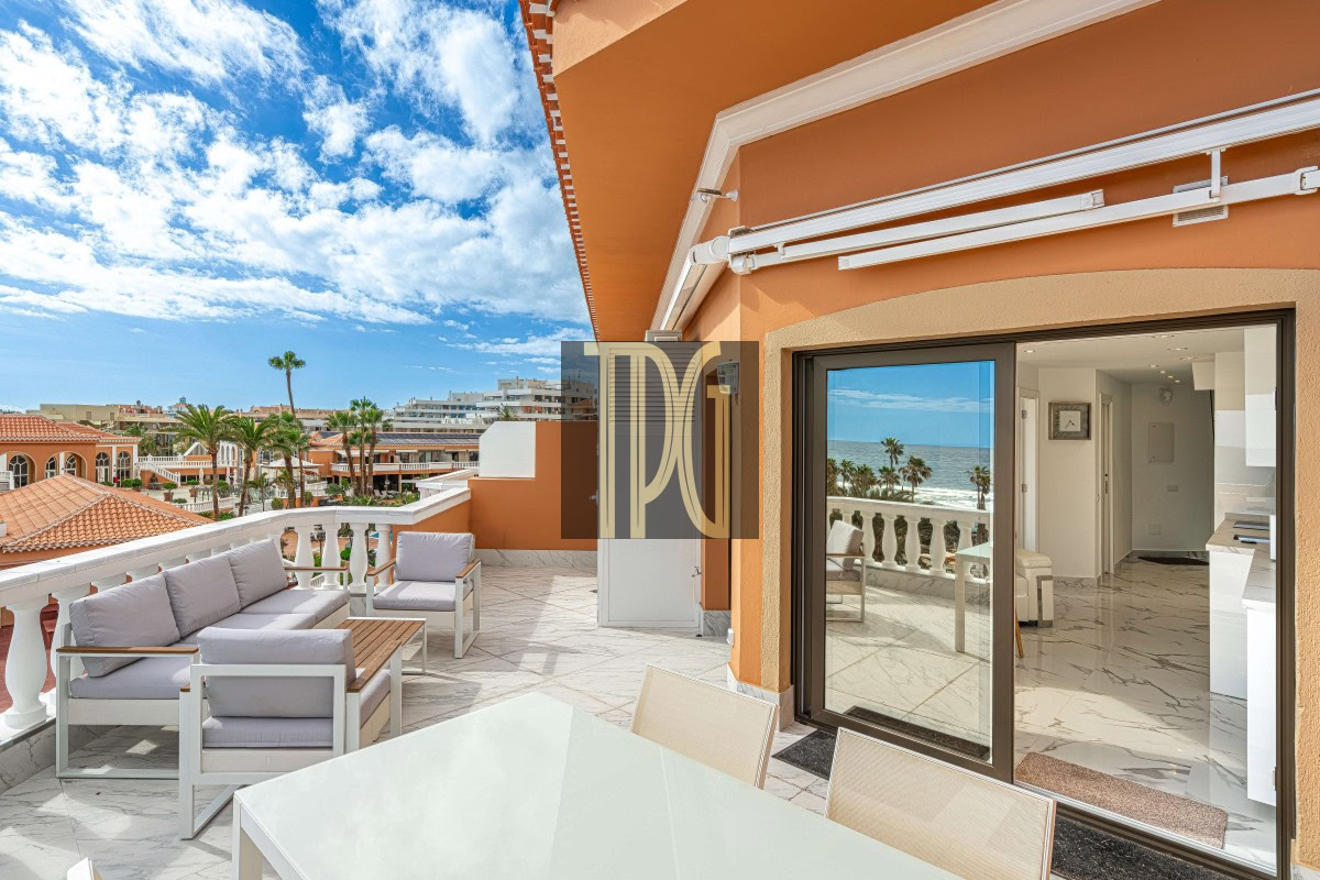 Terrace with white outdoor seating, glass table, and ocean view; orange building exterior and sliding glass doors to the indoor kitchen.