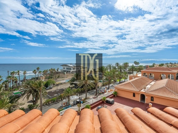 Coastal town view with terracotta rooftops, palm trees, and the blue ocean under a partly cloudy sky.