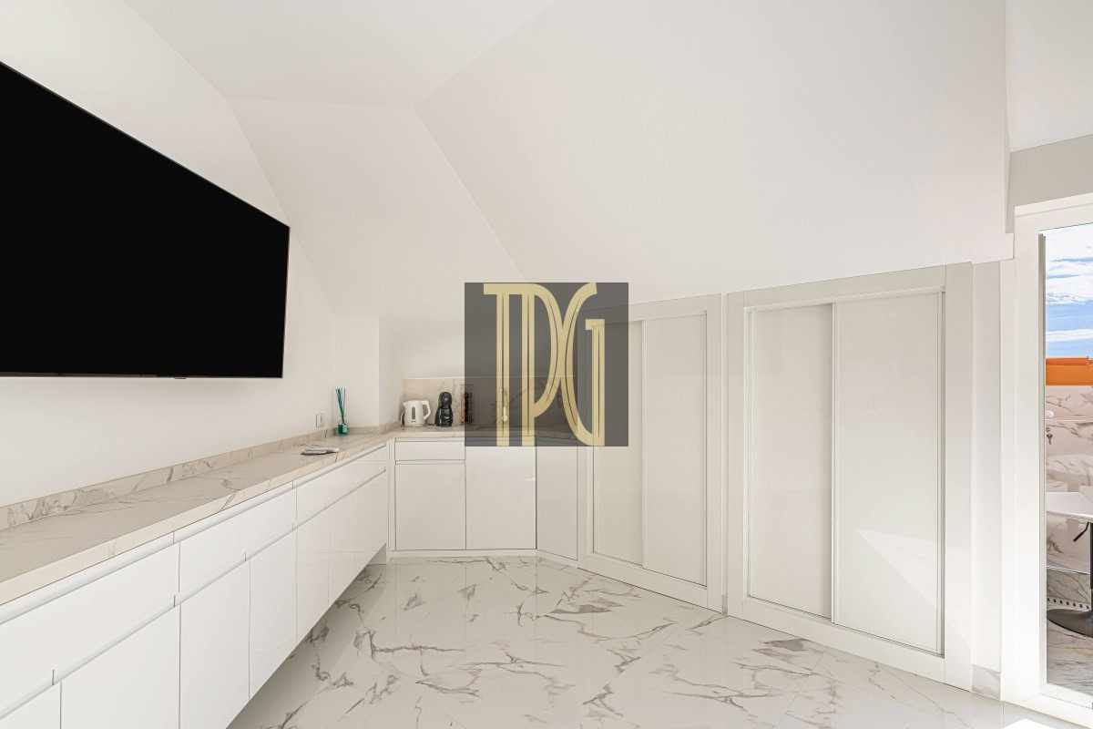 White minimalist kitchen with marble countertops and floors, large wall-mounted TV, and a glass door to the outdoor balcony, high ceilings.