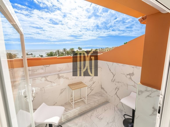 Small marble-walled balcony overlooking the ocean with palm trees and blue sky; two white chairs and a small wooden table against orange walls.