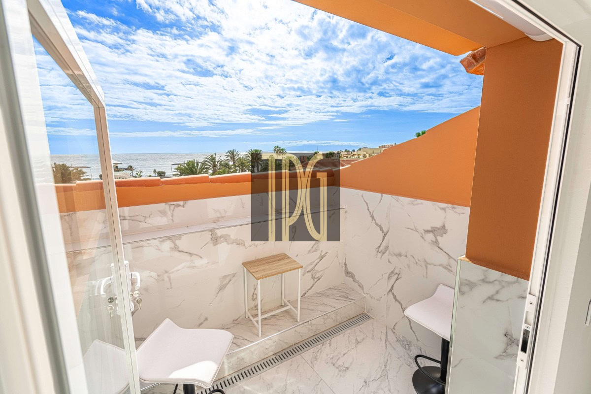 Small marble-walled balcony overlooking the ocean with palm trees and blue sky; two white chairs and a small wooden table against orange walls.
