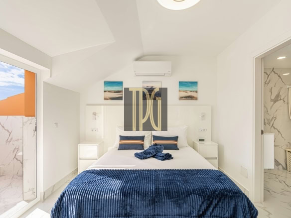 Bright modern bedroom with white walls, two nightstands, and a bed dressed in a navy blue throw blanket and pillows, plus a balcony door to the outside.