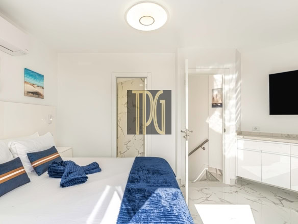 Bright modern white bedroom with a large bed, white bedding, blue accent pillows and throw, marble floor, and a wall-mounted TV on the right side.