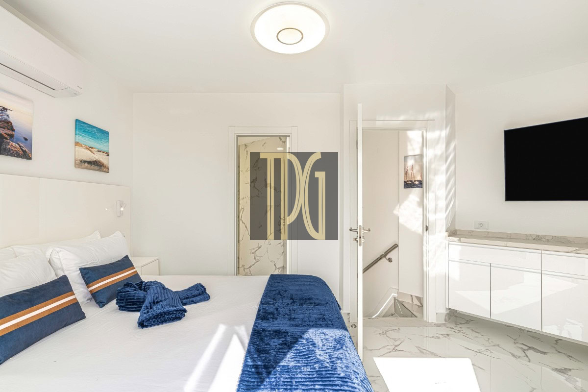 Bright modern white bedroom with a large bed, white bedding, blue accent pillows and throw, marble floor, and a wall-mounted TV on the right side.