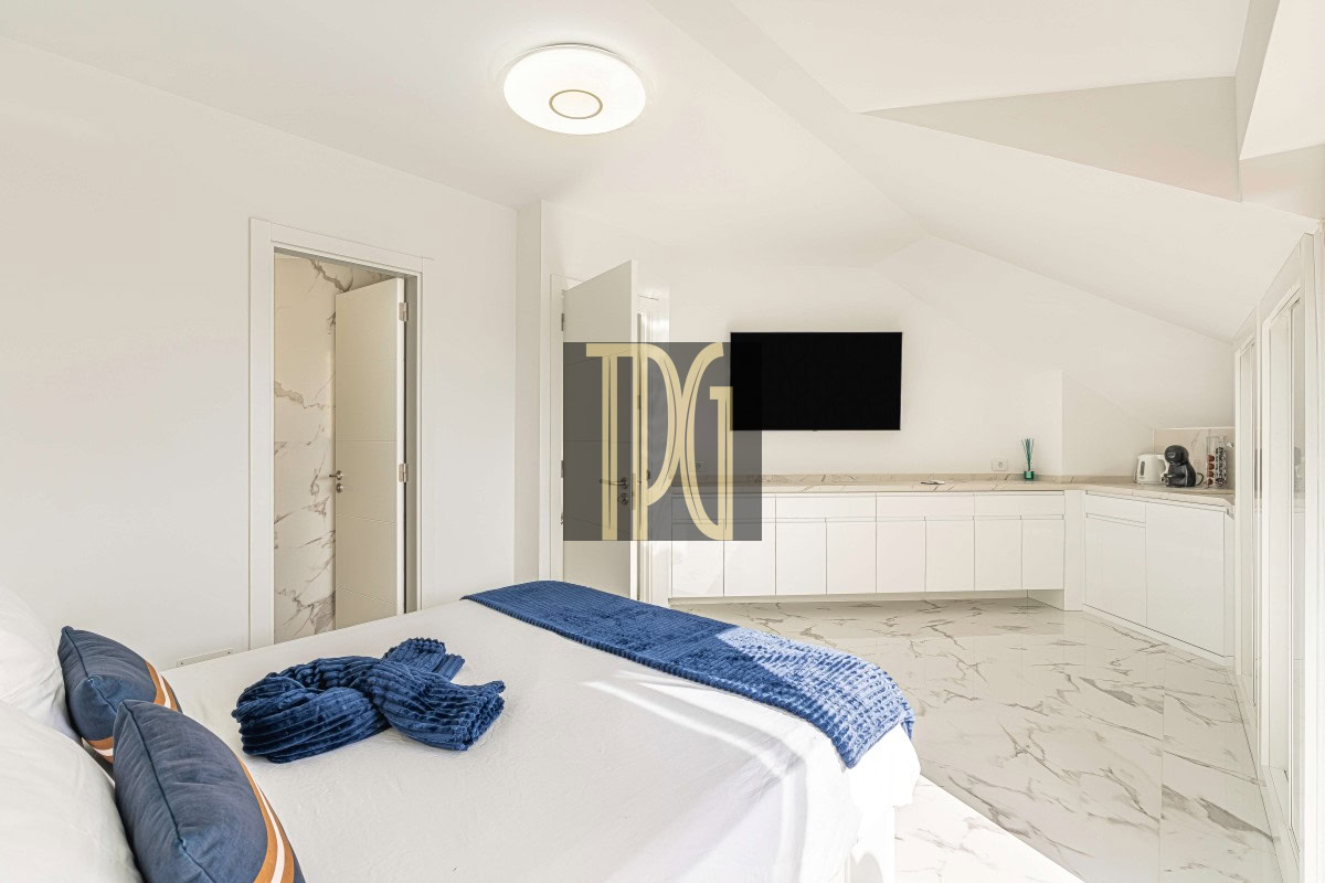 Bright white, minimalist bedroom with a bed in the foreground, blue accent pillows and throw, and a marble-tiled floor; a kitchenette runs along the back wall with a wall TV above.
