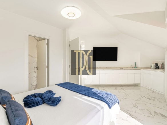 Bright white minimalist bedroom with a bed in the foreground, blue accents, and a marble-like floor, plus a wall-mounted TV over a white cabinet kitchenette.