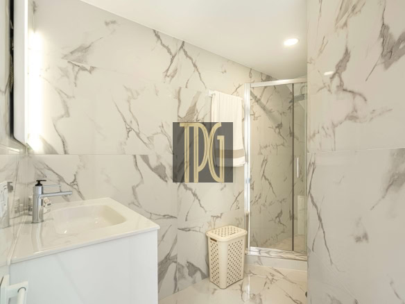 White marble bathroom with a rectangular sink and chrome faucet, towel rack, glass shower enclosure, and a beige laundry basket on the floor.