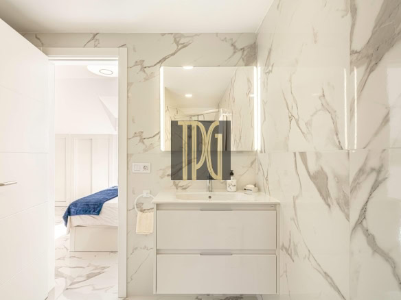 Bright marble-clad bathroom with a white vanity and backlit mirror; doorway reveals a bedroom with a bed and blue throw.