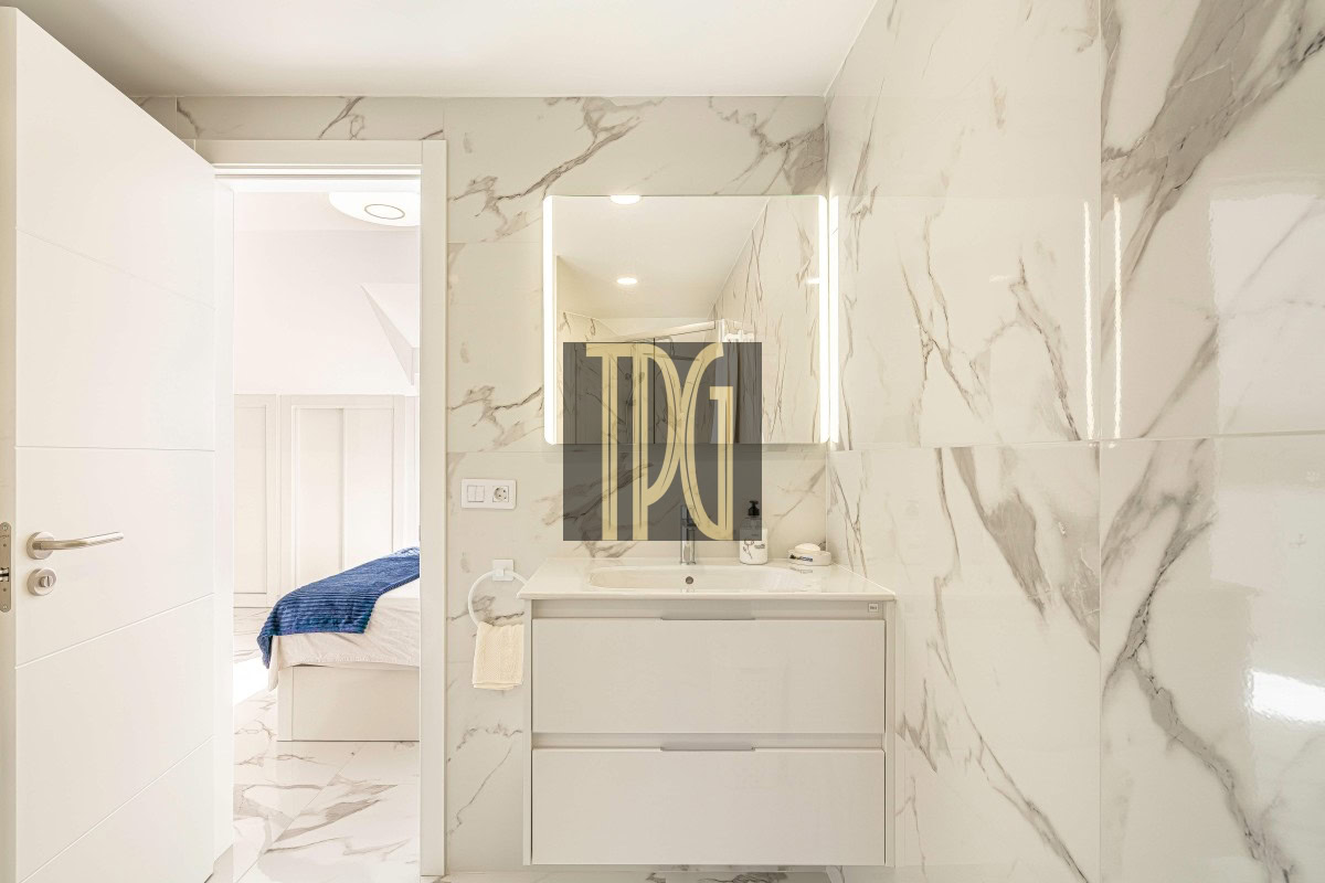 Bright marble-clad bathroom with a white vanity and backlit mirror; doorway reveals a bedroom with a bed and blue throw.