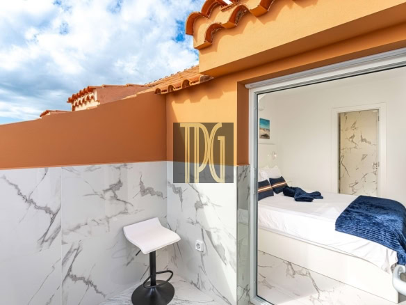 Terrace with orange walls and marble-tiled lower wall, a small white bar stool, and a view into a white bedroom with a navy blue throw on the bed through an open glass door.