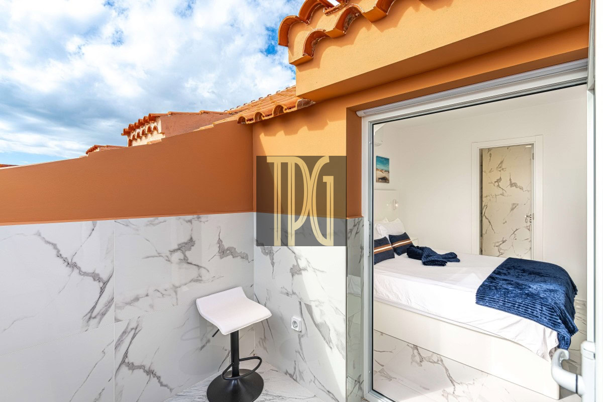 Terrace with orange walls and marble-tiled lower wall, a small white bar stool, and a view into a white bedroom with a navy blue throw on the bed through an open glass door.