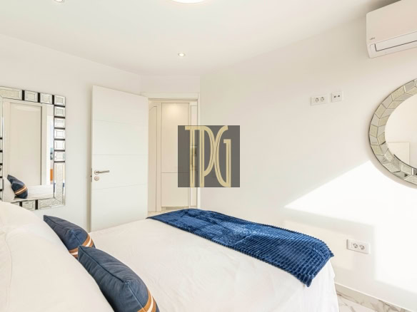 Bright minimalist white bedroom with a bed, blue knit throw, and navy pillows; a square mirrored wall and a round mosaic mirror decorate the walls.