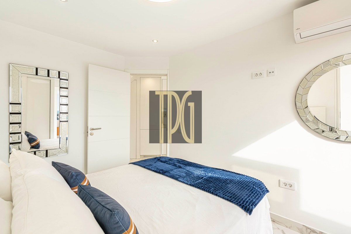Bright minimalist white bedroom with a bed, blue knit throw, and navy pillows; a square mirrored wall and a round mosaic mirror decorate the walls.