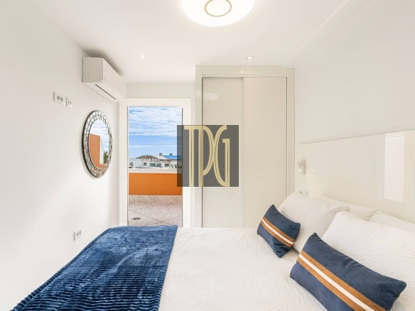 Bright white bedroom with a large bed, blue throw, and navy pillows with orange stripes facing a balcony door outside.