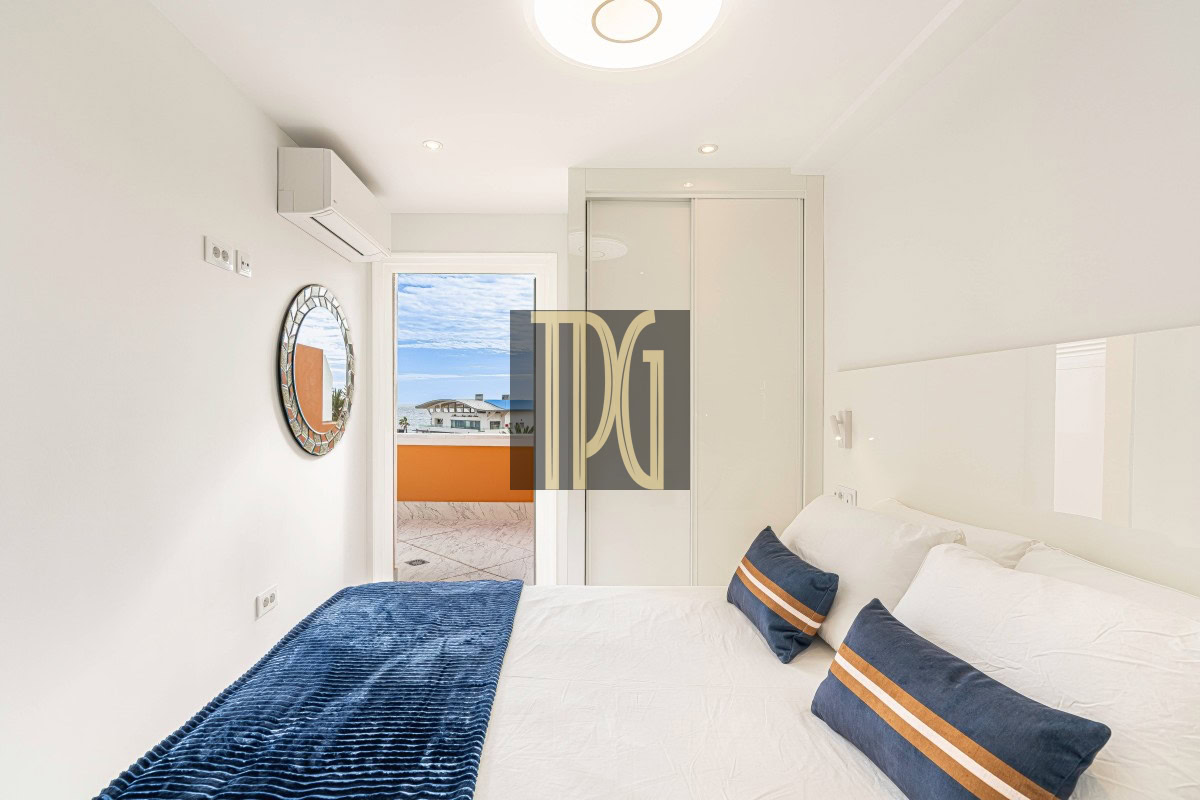 Bright white bedroom with a large bed, blue throw, and navy pillows with orange stripes facing a balcony door outside.