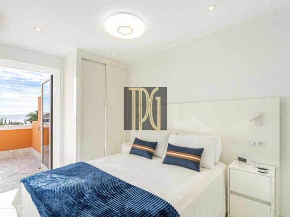 Bright white bedroom with a balcony door open to an orange railing and ocean view, bed with blue pillows and throw.