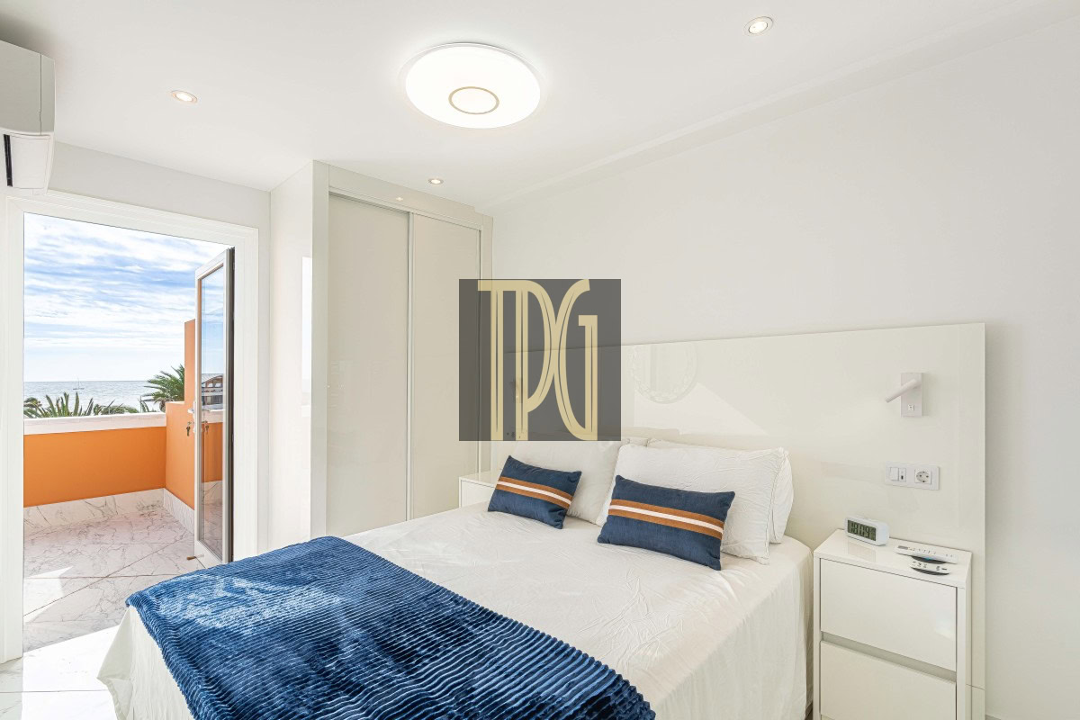 Bright white bedroom with a balcony door open to an orange railing and ocean view, bed with blue pillows and throw.