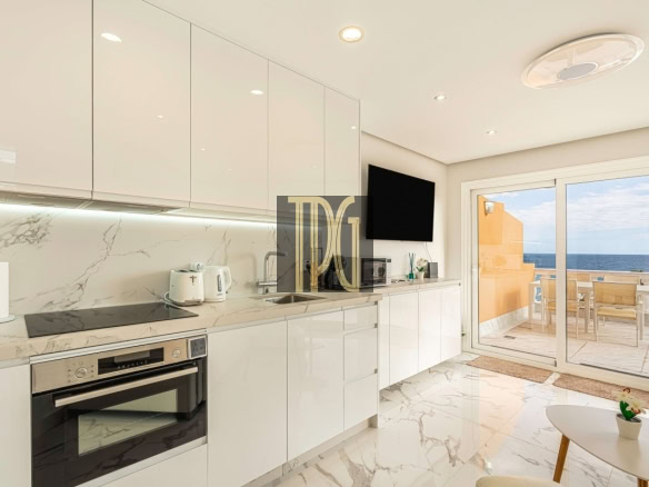 Bright white kitchen with marble countertops and cabinets, built-in oven, and a balcony door showing the ocean view.